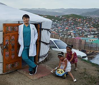 A doctor in Ulaanbaatar, Mongolia, poses for the camera, outside her clinic on the outskirts of Ulaanbaatar and two little girls play on a toy car in the foreground. © Novartis.